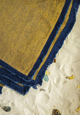 The Riviera Blanket is made of linen in golden sand brown, its soft double-sided design adorned with a fun cotton fringe in deep blue.