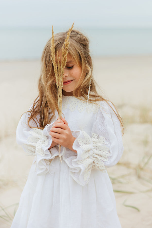 Exquisite soft linen knee-length beach dress with majestic cotton lace details, bishop sleeves, a self-tie belt, and a covered button fastening in the back.